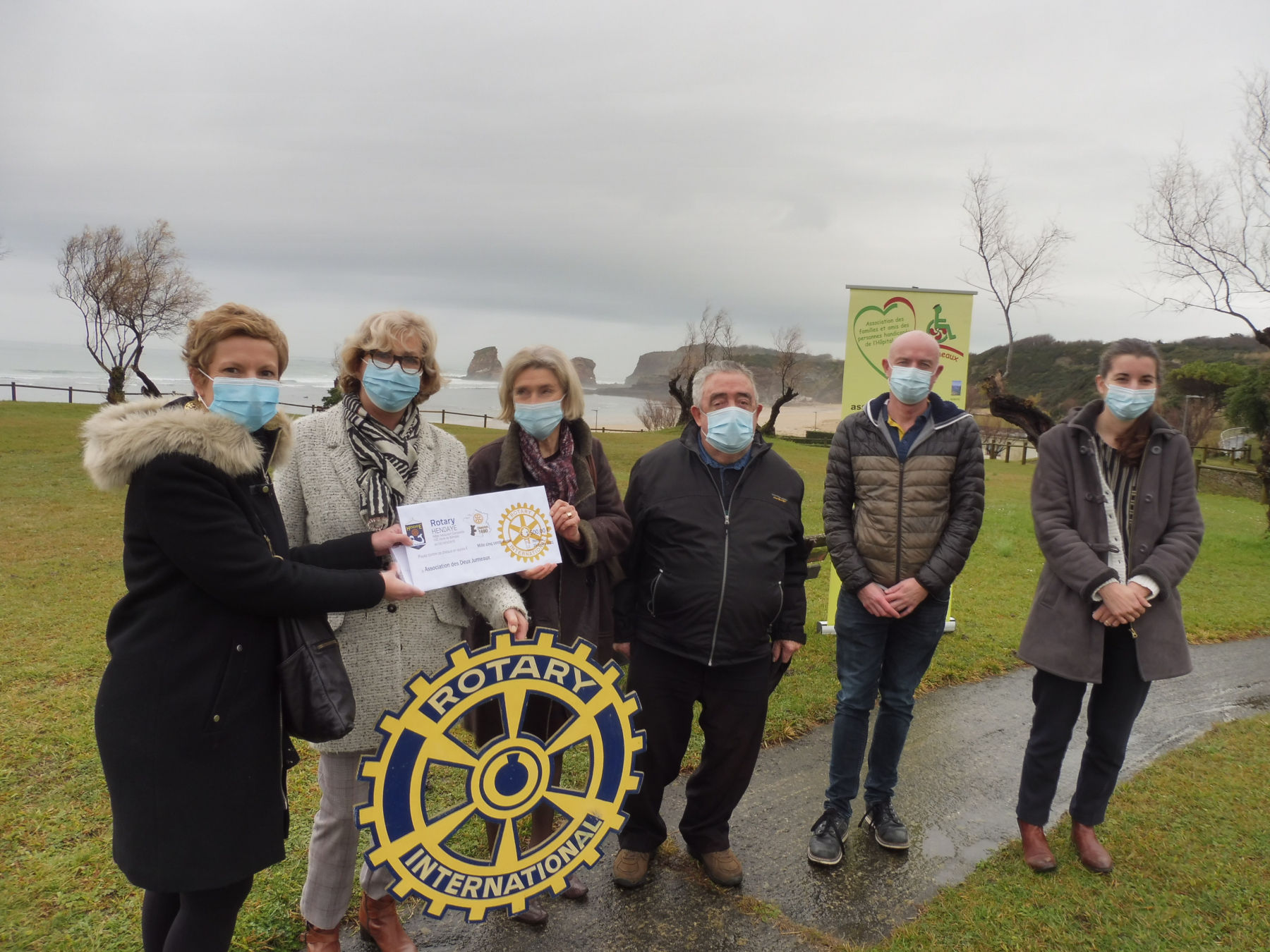 photo officielle de la remise du chèque du Rotary club d'Hendaye à l'association les deux jumeaux à l'hôpital marin de hendaye avec en fond les rochers "les deux jumeaux" symboles de l'association. Temps gris.