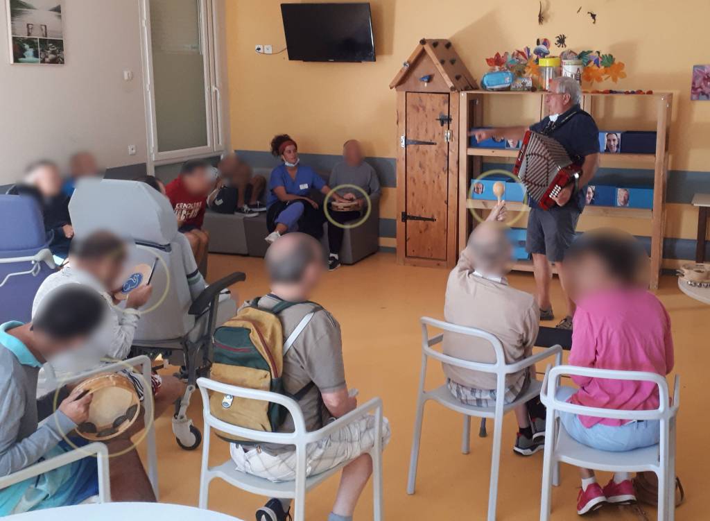 les patients sont de dos, beaucoup ont un instrument de percussion dans les mains et Mattin au centre les fait jouer à tour de rôle, il est avec son triki tritcha (accordeon), le tout dans un lieu de vie couleur beige