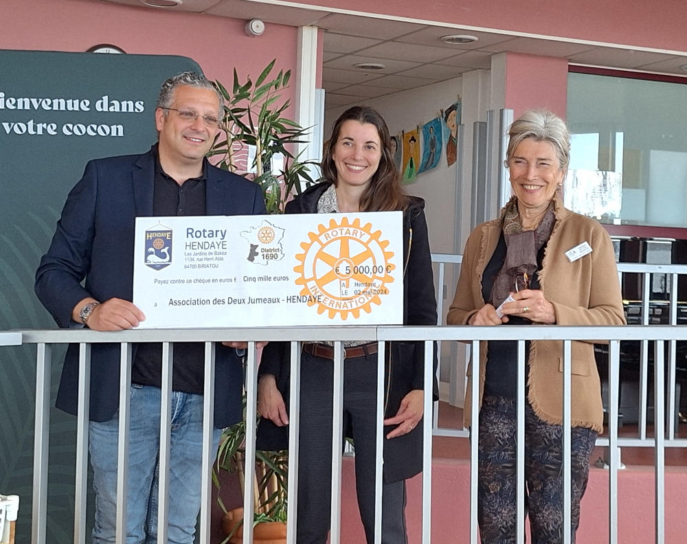 en intérieur de gauche à droite : Benjamin Frischer (rotary), Claire Berriot (directrice adjointe de l'hôpital marin de hendaye) et Ghislaine Bellan Roquette, présidente de l'association Les Deux Jumeaux. Grande affiche représentant un chèque de 5000 euros