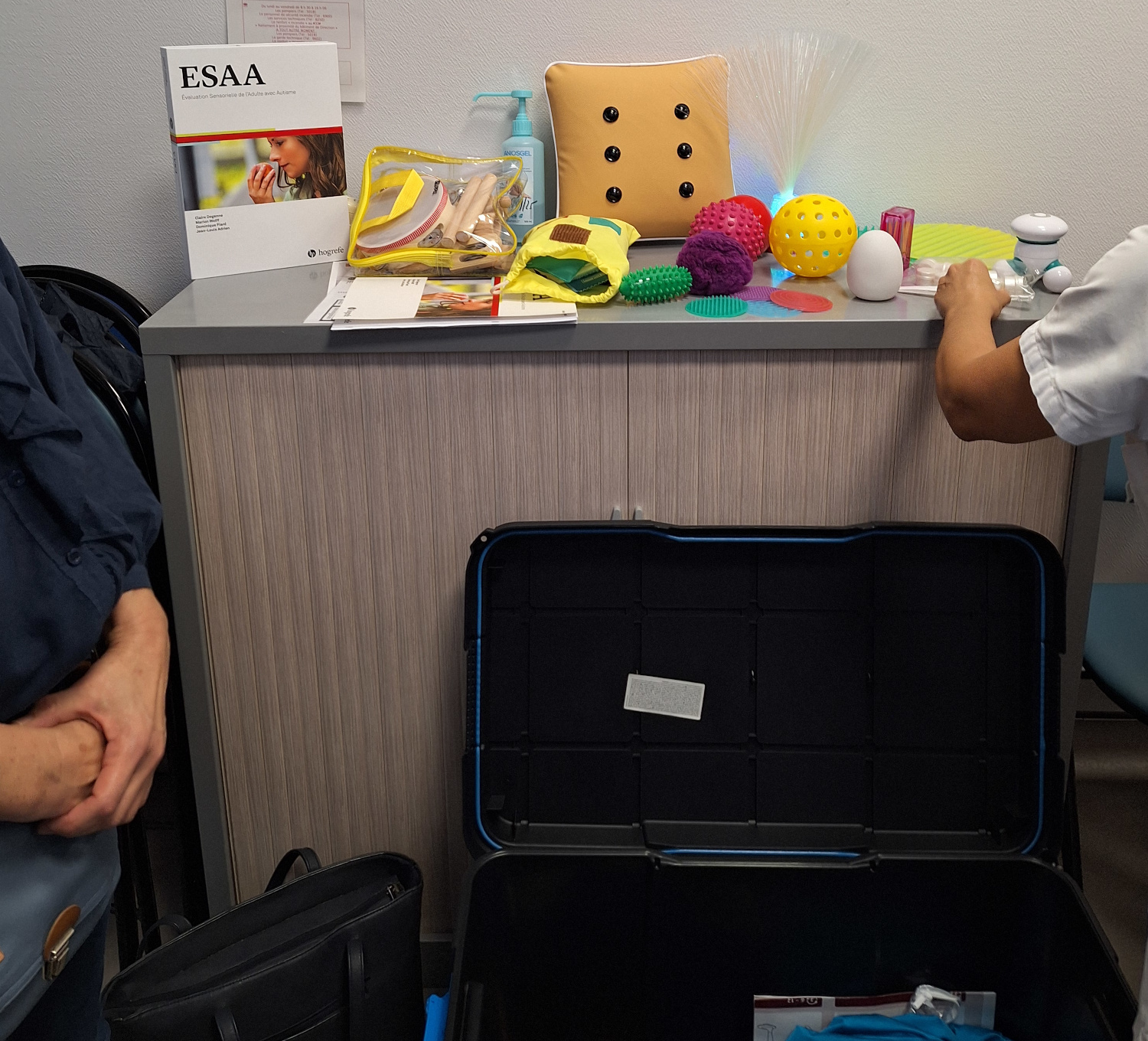 la mallette ouverte en bas puis en haut quelques objets : le guide d'utilisation ESAA et des éléments tactiles : balles, odorants : pots de senteurs, lumineux...