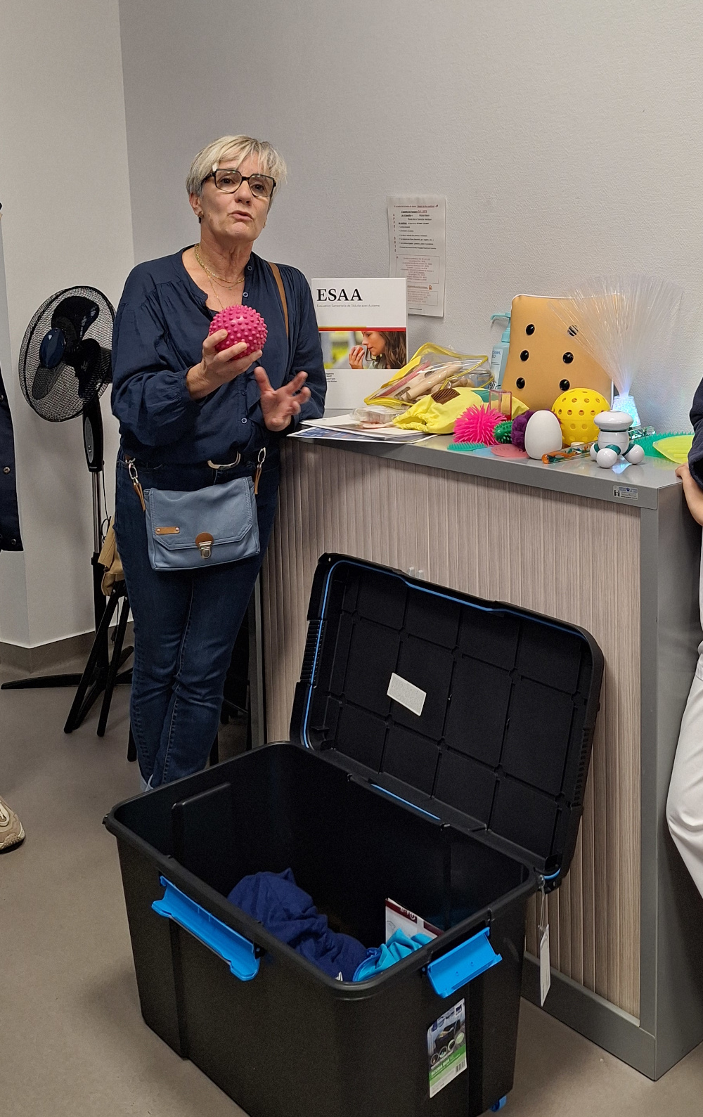 la psychomotricienne tient une balle rouge dans la main en donnant des explications et à sa gauche divers éléments de la mallette sensorielle