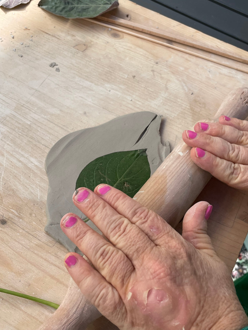 des mains sur un rouleau de bois qui applatit une vraie feuille sur de la glaise